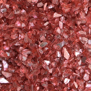 May include: Close-up of a pile of crushed, reflective, red glass pieces. The fragments vary in size and shape, catching the light and creating a sparkling effect. The overall texture is rough and irregular, with a vibrant, ruby-like color.