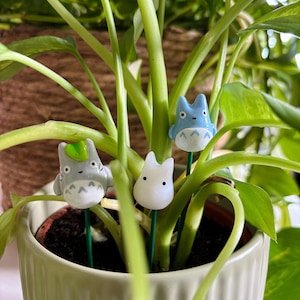 Puede incluir: Tres marcadores de plantas de cerámica con forma de personajes de dibujos animados. Los personajes son azules, blancos y grises. Los marcadores se insertan en una planta verde en maceta.