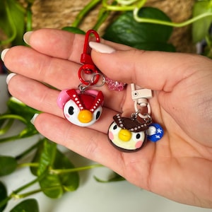 Peut inclure: Deux porte-clés faits à la main en forme de hibou. L'un est rouge et rose avec un bec jaune, l'autre est marron et crème avec un bec jaune. Les deux ont un petit fermoir en métal pour les attacher aux clés ou aux sacs. Les porte-clés sont tenus dans une main.