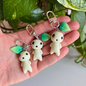 Puede incluir: Tres llaveros con una pequeña figura blanquecina con ojos negros y un adorno de hoja. Los llaveros tienen cierres plateados. Las figuras se sostienen en una mano, con follaje verde en el fondo.