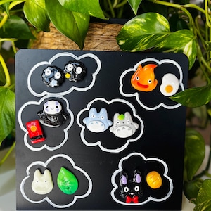May include: A black square with white cloud outlines, featuring colorful character magnets. The magnets include a variety of designs, such as two soot sprites, a smiling spirit, two blue Totoro figures, and a black rabbit with a red bow tie.