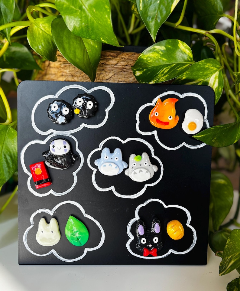May include: A black square with white cloud outlines, featuring colorful character magnets. The magnets include a variety of designs, such as two soot sprites, a smiling spirit, two blue Totoro figures, and a black rabbit with a red bow tie.