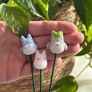 May include: Three ceramic figurines of characters from the animated film My Neighbor Totoro. The figurines are attached to green metal stems. The figurines are light blue, gray, and white.