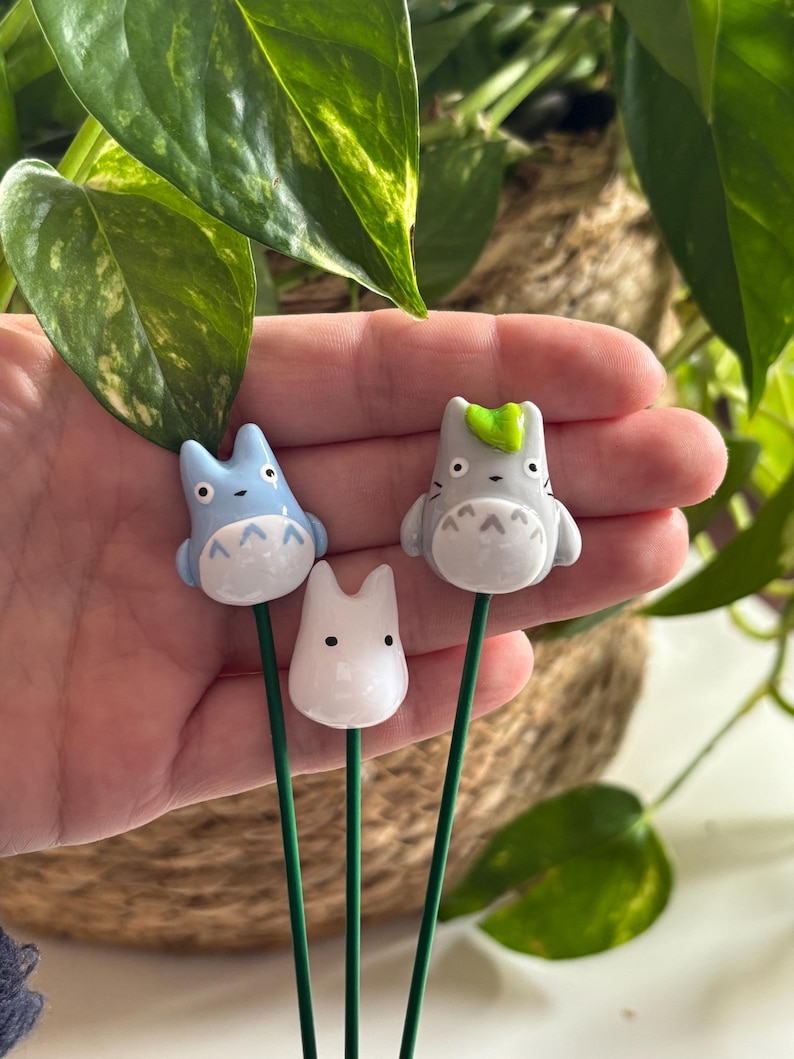 May include: Three ceramic figurines of characters from the animated film My Neighbor Totoro. The figurines are attached to green metal stems. The figurines are light blue, gray, and white.