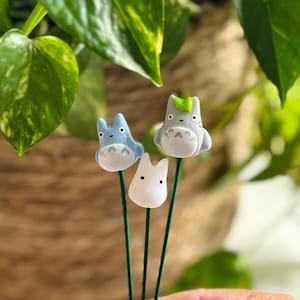 May include: Three ceramic figurines of characters from the animated film My Neighbor Totoro. The figurines are attached to green metal stems. The figurines are white, light blue, and gray with black eyes and a light blue nose.