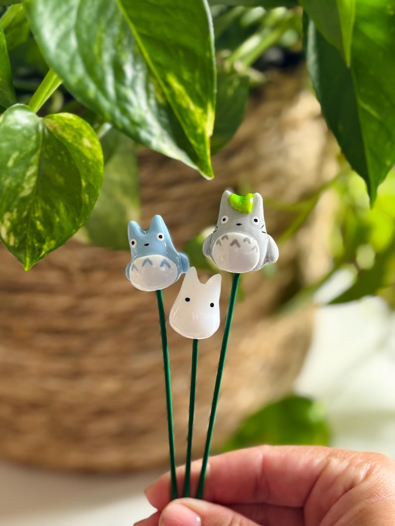 May include: Three ceramic figurines of characters from the animated film My Neighbor Totoro. The figurines are attached to green metal stems. The figurines are white, light blue, and gray with black eyes and a light blue nose.