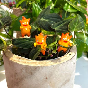 May include: A concrete planter filled with a lush green plant, adorned with three small, orange, cartoon-like figures. The figures have a flame-like head, black eyes, and a red mouth. The plant has large, dark green leaves.