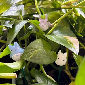 May include: Three small ceramic figurines of a cartoon character with different colored fur. The figurines are light gray, light blue, and white. The figurines are placed in a pot of green leafy plants.