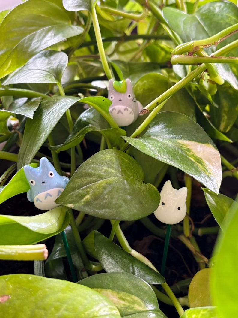 May include: Three small ceramic figurines of a cartoon character with different colored fur. The figurines are light gray, light blue, and white. The figurines are placed in a pot of green leafy plants.