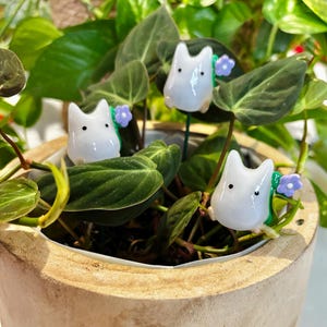 May include: A close-up of a houseplant in a wooden pot, decorated with three white, cartoon figurines. Each figurine has a small purple flower. The plant has green leaves, and the pot is light brown.