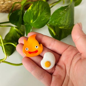 May include: Two handmade, colorful resin charms. One is a smiling orange flame with black eyes and a red mouth. The other is a white egg with a yellow yolk. The charms are held in a hand.