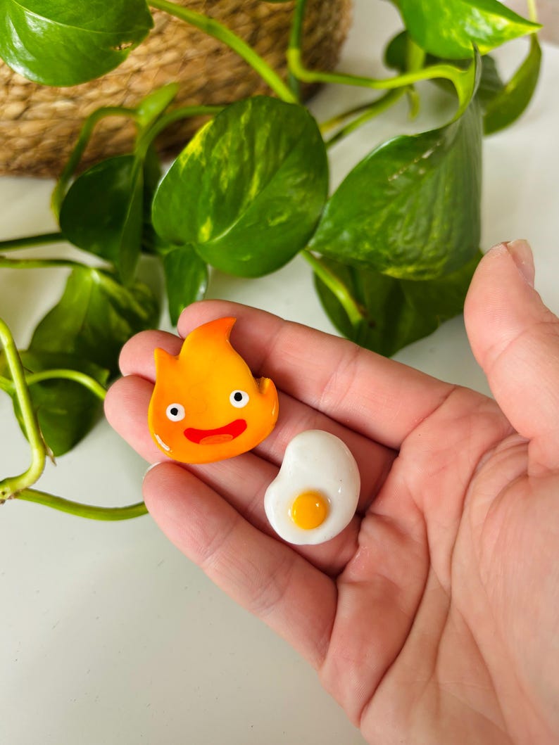 May include: Two handmade, colorful resin charms. One is a smiling orange flame with black eyes and a red mouth. The other is a white egg with a yellow yolk. The charms are held in a hand.