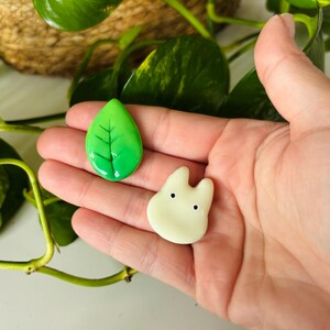 May include: Two handmade resin charms: a green leaf with black veins and a cream-colored, cartoon-like character with two black dots for eyes. The charms are held in a hand, with green plant leaves and a woven basket in the background.