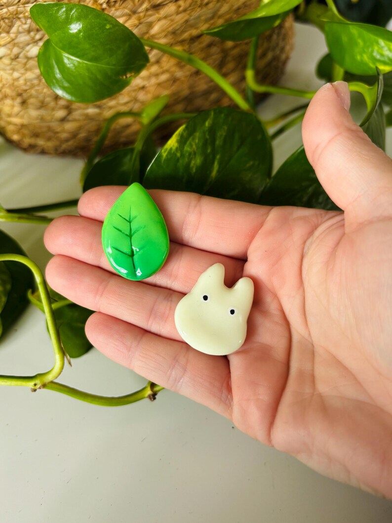 May include: Two handmade resin charms: a green leaf with black veins and a cream-colored, cartoon-like character with two black dots for eyes. The charms are held in a hand, with green plant leaves and a woven basket in the background.