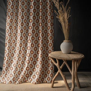 May include: Cream-coloured curtain featuring a repeating pattern of brown and orange circles linked by vertical lines. A small, round, woven side table with a grey vase holding dried plants is to the right. The background is dark.
