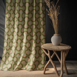 May include: A green and peach geometric patterned curtain hangs next to a small, round, woven side table. A gray vase with dried grasses sits on the table. The curtain's design features interlocking circles.