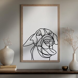 May include: A framed art print featuring a minimalist line drawing of a dog's face. The black line art is set against a white background, encased in a light brown wooden frame. The artwork is displayed on a dark brown table.