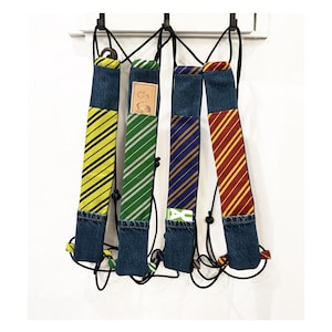 May include: Four colorful, striped tie-themed backpack straps with denim bases hang against a white background. The straps feature yellow and black, green and white, blue and gold, and red and gold stripes. Each strap has a black cord and a small loop.