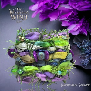 May include: A cuff bracelet featuring a complex weave of green thread, purple, lime green, and olive fabric strips, accented with purple beads, metallic fibers, and a central iridescent stone. The text reads The Whispering Wind Cuff & Woodland Grape.