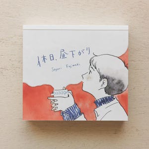 May include: A square notepad with an illustration of a person enjoying a hot beverage. The artwork features a person with short hair, wearing a blue and white striped sweater, and holding a cup. The background is red and white, with Japanese text and the name "Sayuri Fujimaki".