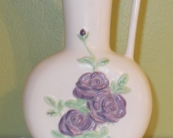 May include: A white ceramic pitcher with a handle and a floral design. The pitcher features three purple roses with green leaves.
