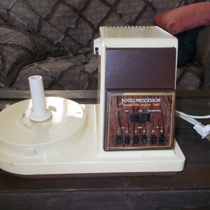May include: A white and brown food processor with a wooden-grain design on the control panel. The panel has a dial with "On", "Off", and "Momentary" settings, and a row of six buttons numbered 1 through 6. The text "Food Processor" and "Hampton Beach" is printed on the panel.