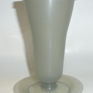 May include: A tall, translucent gray plastic tumbler with a flared top and a matching circular base stand. The container features a smooth, matte finish and is topped with a solid, opaque white circular lid that fits securely over the rim.