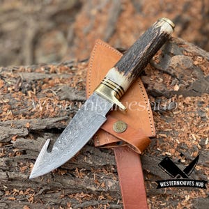 Op de afbeelding: Een Damascus stalen jachtmes met een gebogen lemmet en een geweien handvat rust op een bruine leren schede. Het mes heeft messing accenten en een decoratief embleem op de schede. De afbeelding bevat de tekst "Western Knives Hub".