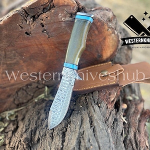 May include: A handmade knife with a patterned blade and a green handle with blue accents. The knife rests on a wooden surface next to a brown leather sheath. The text "Western Knives Hub" is visible.