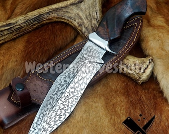 May include: A large, ornate hunting knife with a patterned blade and a dark wooden handle. The knife rests in a brown leather sheath with a stitched border. The image includes the text "WesternKnives Hub".