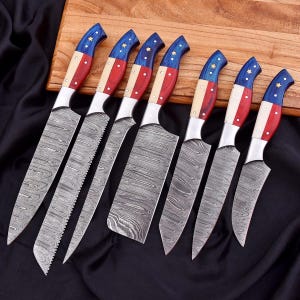 Può includere: Set di sette coltelli da cucina con lame in acciaio decorate e manici rossi, bianchi e blu con stelle. I manici curvi sembrano essere in legno. I coltelli sono disposti su un tagliere di legno.