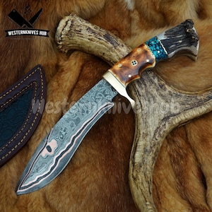 May include: A handcrafted hunting knife with a curved blade, intricate pattern, and a handle made of wood, brass, and turquoise accents. The knife rests on a brown fur surface, with a leather sheath and deer antler in the background. The text "WESTERNKNIVES HUB" is visible.