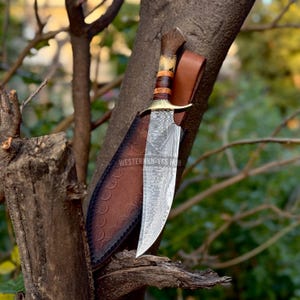 May include: A large hunting knife with a patterned blade and a wooden handle is displayed in a brown leather sheath. The knife has a brass bolster and is partially sheathed. The text "WESTERNKNIVES HUB" is visible on the sheath.