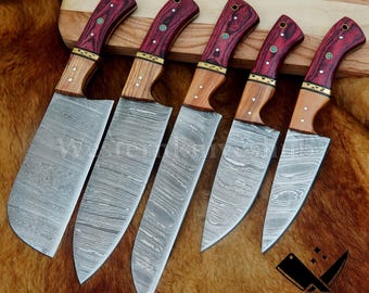 Puede incluir: Un juego de cinco cuchillos de acero de Damasco con mangos de madera. Los mangos combinan madera clara y oscura, acentuados con detalles rojos y dorados. Las hojas presentan un diseño distintivo.