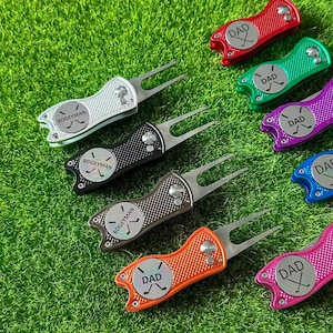 Könnte beinhalten: Eine Sammlung von Golf-Divot-Reparaturwerkzeugen in verschiedenen Farben, darunter Silber, Schwarz, Braun, Orange, Rot, Grün, Lila, Pink und Blau. Jedes Werkzeug hat eine Metallgabel und ein rundes Emblem mit dem Text "BOGEYMAN" oder "DAD".