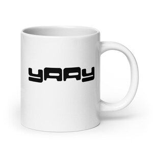 Op de afbeelding: Witte keramische mok met het woord "YAY" in vette, zwarte letters. De mok heeft een gebogen oor en een gladde, glanzende afwerking. Het ontwerp is gecentreerd op de voorkant van de mok.