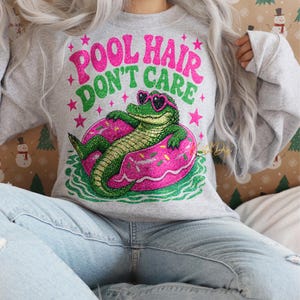 May include: A light grey sweatshirt with the slogan "POOL HAIR DON'T CARE" in pink and green. The graphic shows a green alligator wearing heart-shaped sunglasses, on a pink donut-shaped inflatable. Pink stars and sparkles surround the text.