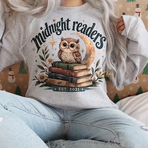 Op de afbeelding: Een lichtgrijs sweatshirt met een grafische afbeelding. Het ontwerp toont een uil op een stapel boeken, een halve maan en de woorden "Midnight Readers" en "EST. 2021". Het ontwerp bevat bloemen en sterren.