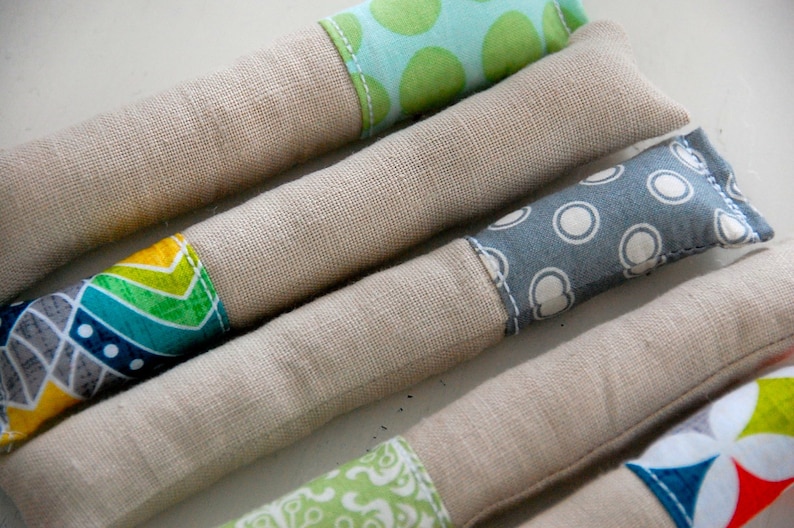 Może przedstawiać: Cztery ręcznie robione zabawki dla kot&oacute;w z kocimiętką, z r&oacute;żnymi wzorzystymi pokrowcami z tkaniny. Zabawki są wykonane z naturalnego lnu i wypełnione kocimiętką. Wzory obejmują zielone i niebieskie kształty geometryczne, niebieskie i białe kropki oraz zielone i ż&oacute;łte kształty geometryczne.