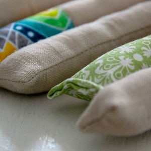 Może przedstawiać: Cztery poduszki do oczu pokryte tkaniną w r&oacute;żnych wzorach. Poduszki są beżowe z r&oacute;żnymi kolorowymi wzorami z przodu. Wzory obejmują zielono-białe kwiaty, niebiesko-białe wzory geometryczne i ż&oacute;łto-zielone wzory geometryczne.