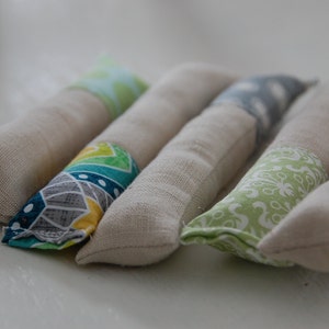 Może przedstawiać: Pięć ręcznie robionych zabawek z kocimiętką wykonanych z naturalnego materiału z r&oacute;żnymi kolorowymi wzorami. Zabawki mają kształt długich cylindr&oacute;w i są wypełnione kocimiętką.