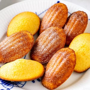 Puede incluir: Un plato de madeleines doradas, un clásico pastel francés en forma de concha, espolvoreadas con azúcar glas. Los pasteles están dispuestos en un plato blanco con un diseño floral azul.