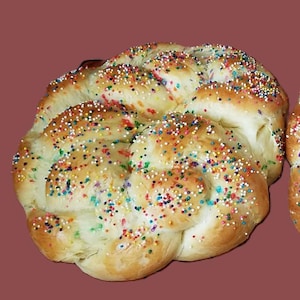 Puede incluir: Una trenza de pan dorada, cubierta con una generosa capa de chispas de colores. El pan tiene una textura suave y esponjosa y es probablemente un pan dulce, perfecto para una ocasión festiva.
