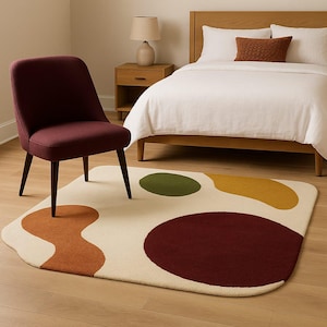 May include: A burgundy upholstered chair with dark wooden legs sits on a cream-colored rug with abstract shapes in rust, olive green, and mustard yellow. A bed with white bedding and a wooden frame is in the background.