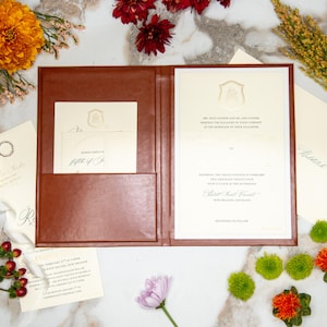 Puede incluir: Un juego de invitaciones de boda de cuero marrón, abierto para revelar una invitación color crema con letras doradas y un bolsillo para tarjetas de respuesta. Acentos florales rodean la invitación, incluyendo flores naranjas y burdeos.