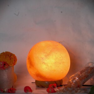 Puede incluir: Una lámpara esférica de sal del Himalaya emite una cálida luz naranja. La lámpara se encuentra sobre una base de madera, con un pequeño jarrón de flores y un tarro de cristales de sal derramados cerca. El tema general es la decoración natural del hogar y el bienestar.