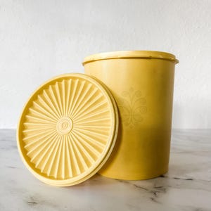 Puede incluir: Un recipiente de plástico amarillo vintage con una tapa a juego. La tapa es redonda con un diseño de rayos de sol. El recipiente tiene un diseño floral en el lateral. El recipiente y la tapa están hechos de un material plástico duradero.