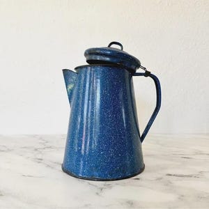 Könnte beinhalten: Eine blaue, gesprenkelte Emaille-Kaffeekanne mit Deckel. Die Kanne hat einen Griff und einen Ausguss. Der Deckel ist leicht geöffnet. Die Kanne steht auf einer Marmoroberfläche.