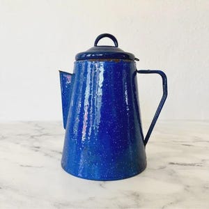 Könnte beinhalten: Eine blaue Emaille-Kaffeekanne im Vintage-Stil mit Deckel und Griff. Die Kanne hat ein gesprenkeltes Muster und einen Ausguss zum Ausgießen. Der Griff ist dunkelblau. Die Kanne steht auf einer Marmoroberfläche.