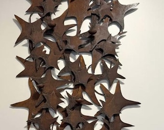 May include: A wall-mounted metal sculpture composed of numerous star-shaped elements. The stars have a rustic, weathered appearance with a dark brown, almost rusty color. The arrangement is dense, creating a textured, three-dimensional effect.